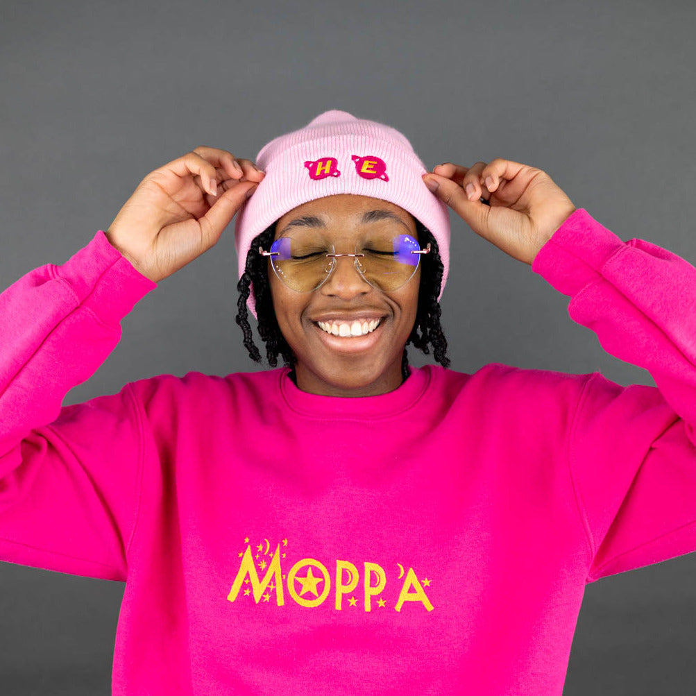 Person wearing a bright pink sweatshirt with "moppa" gold text and pink beanie with "he" gold text, against a gray background